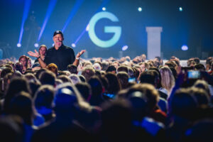 A man in a black shirt and cap gesticulates enthusiastically in front of a large audience. In the background, a large "G" lights up on a stage, surrounded by blue lighting effects.