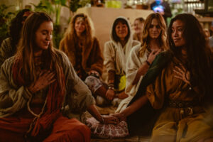 Two people are sitting in the foreground, holding hands and placing their other hand on their heart. Several people in a relaxed atmosphere in the background, decorated with plants and warm light.