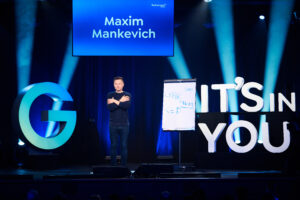 Ein Mann steht auf einer B&uuml;hne mit verschr&auml;nkten Armen. Rechts von ihm steht eine beschriebene Staffelei, links ein gro&szlig;es &bdquo;G&ldquo;. Im Hintergrund leuchtet ein Schild mit &bdquo;Maxim Mankevich&ldquo;. Daneben steht IT'S IN YOU.